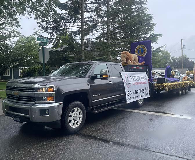 A gray Chevrolet pickup truck pulling a colorful float. The float had decorations and a big sign that said "Lions Club." There was another sign on the side of the truck with phone numbers and information. On the float, there was a big mascot that looked like a lion. - Taylor Made Truck Driving School