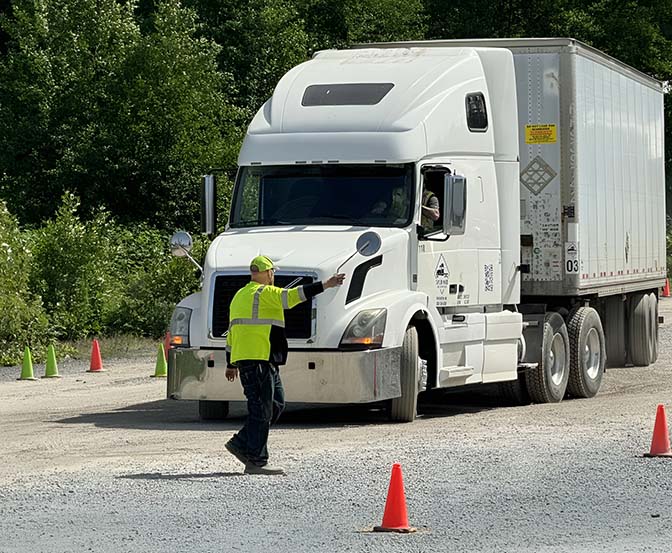 A person wearing a bright, easy-to-see vest and hat is waving to a big white truck. The truck is driving on a rocky road with orange cones all around, and there are trees and bushes in the background. - Taylor Made Truck Driving School