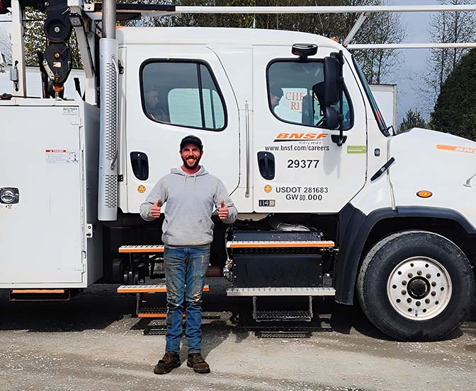 A person wearing a gray hoodie and jeans is standing and smiling in front of a BNSF work truck. They are giving a thumbs-up with both hands. You can see the truck's logo and some numbers on its side. - Taylor Made Truck Driving School