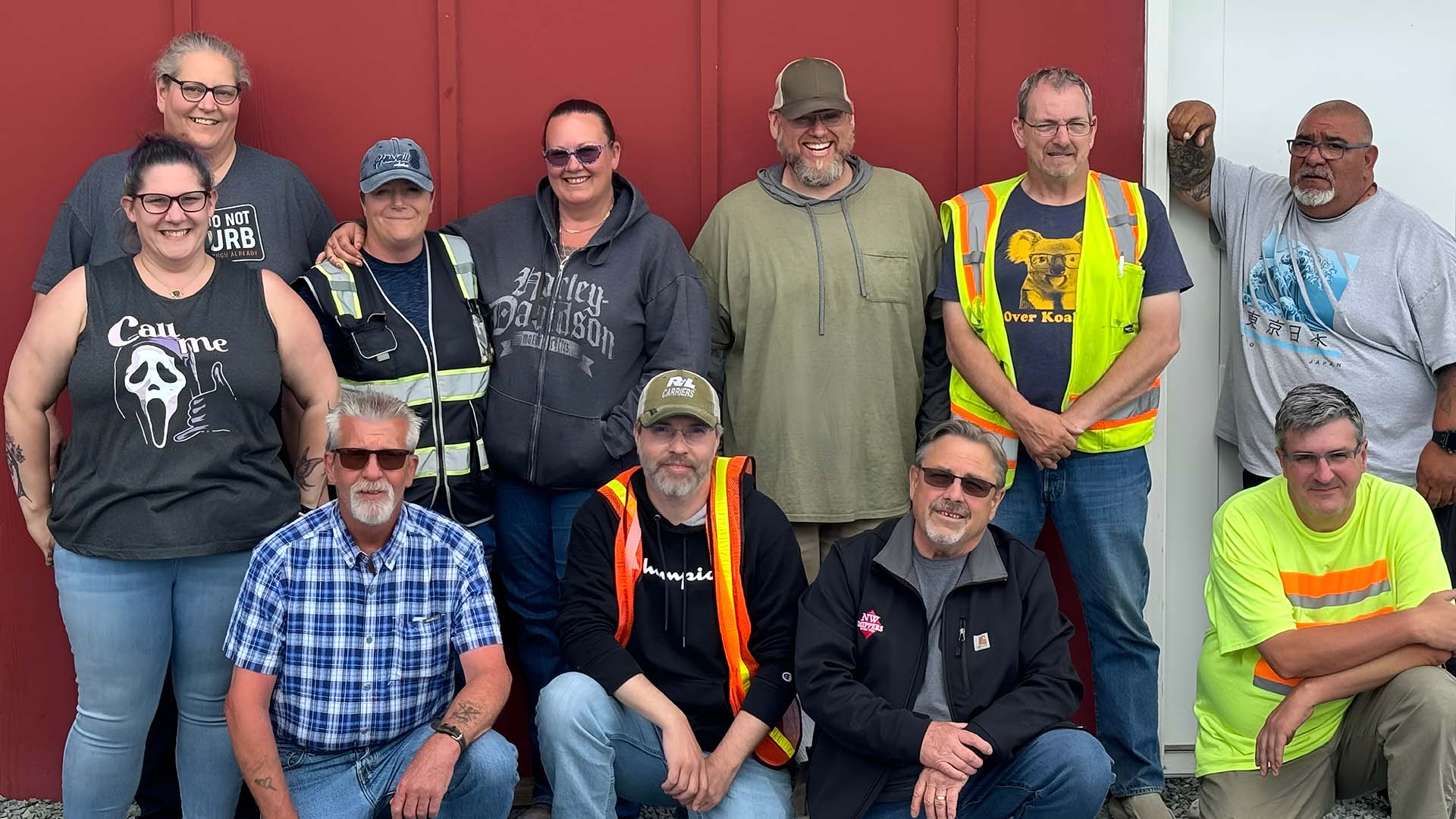 Truck driving school instructors. They are in front of a red and white wall. Some people are standing up, while others are squatting or sitting on the ground. - Taylor Made Truck Driving School